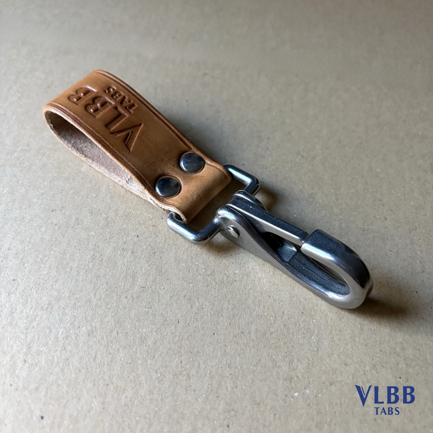 Natural leather belt loop with a stainless steel snap hook fixed by two rivets, VLBB TABS stamped on the leather, on brown cardboard